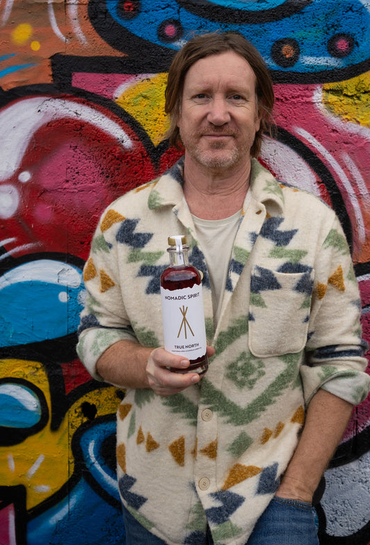 A man against a bright painted wall, holding a bottle of True North, relaxed and looking straight at the camera